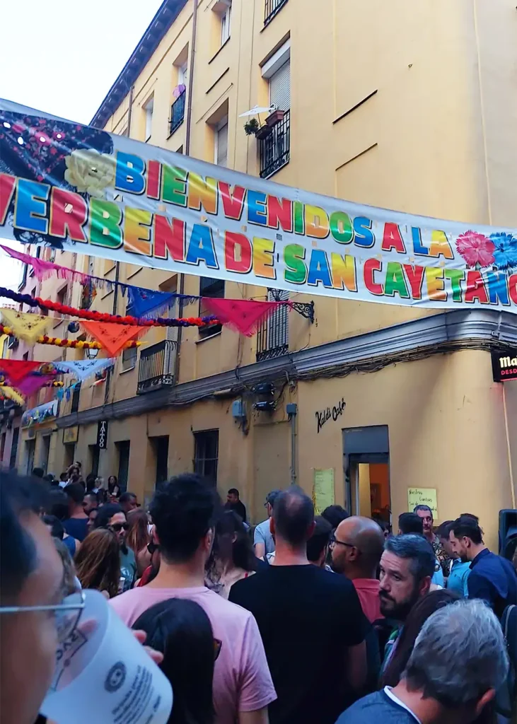 Faixa colorida com a frase "Bienvenidos a la Verbena de San Cayetano" estendida sobre uma rua lotada de pessoas durante as festas de agosto em Madrid.