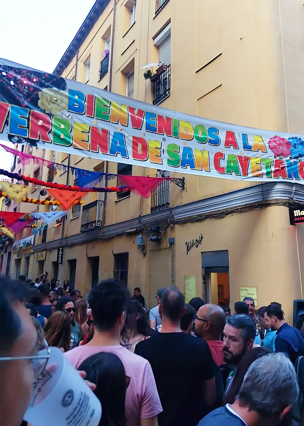 Faixa colorida com a frase "Bienvenidos a la Verbena de San Cayetano" estendida sobre uma rua lotada de pessoas durante as festas de agosto em Madrid.