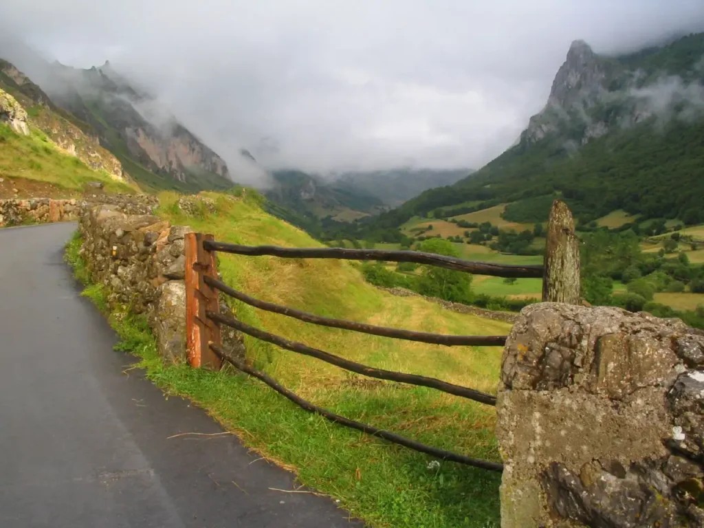 Estrada asfaltada ao lado de uma cerca de madeira, com vista para os vales verdes de Somiedo cobertos por neblina.