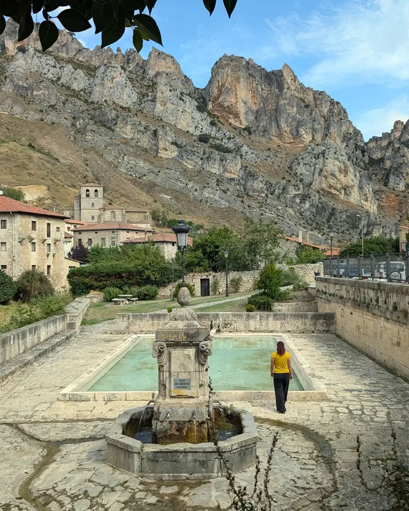 Mulher de costas, vestindo blusa amarela, em frente ao lavadouro público de Pancorbo, com as casas do vilarejo e os rochedos do desfiladeiro ao fundo.