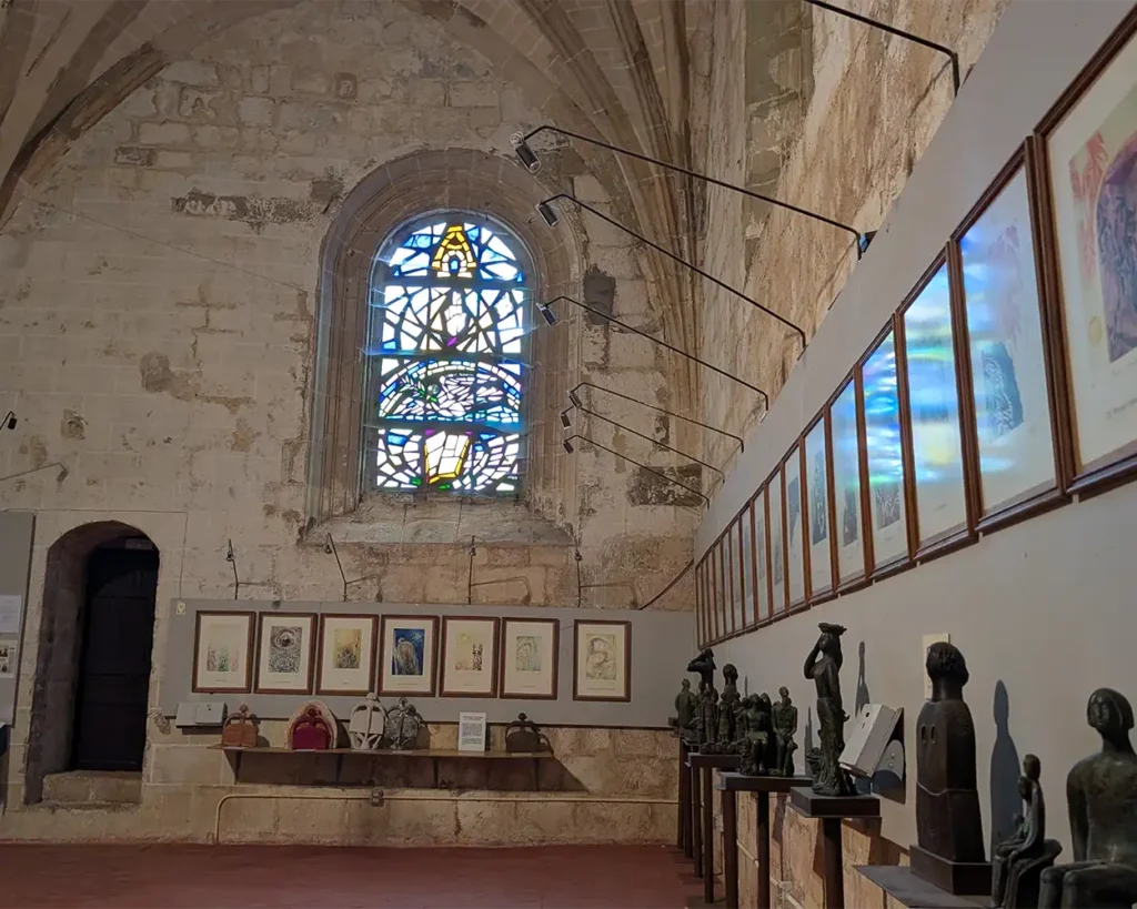 Exposição de arte dentro da Igreja de Santiago em Pancorbo, mostrando esculturas e quadros em uma parede de pedra com um vitral ao fundo.