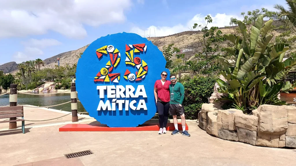 Casal de visitantes posando em frente à placa azul de entrada do parque Terra Mítica, com o logo de 25 anos e a montanha com o letreiro do parque ao fundo.