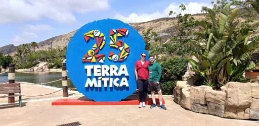 Casal de visitantes posando em frente à placa azul de entrada do parque Terra Mítica, com o logo de 25 anos e a montanha com o letreiro do parque ao fundo.