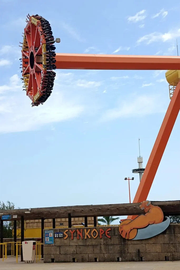 The Synkope thrill ride as seen from its entrance, with the giant pendulum swinging high above and the ride's sign in the foreground.