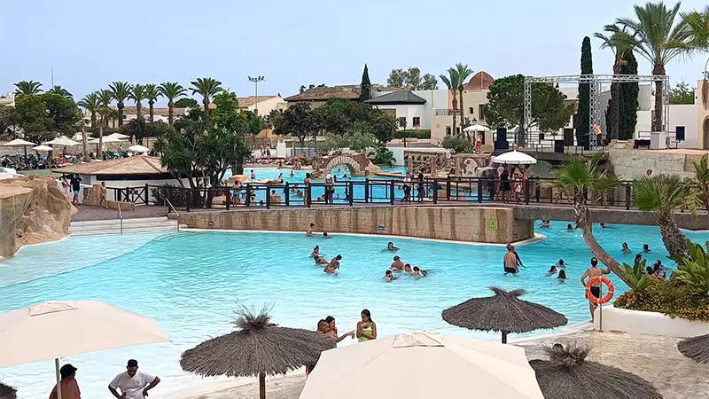 Visitantes se refrescando na piscina principal da Gran Área Spa, no Terra Mítica, com uma ponte e a área de animação ao fundo.