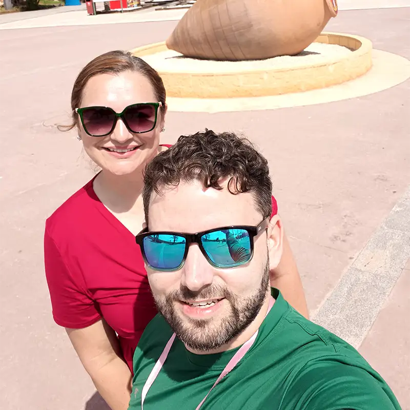 Casal tira uma selfie na área de entrada do Terra Mítica, com palmeiras e uma grande ânfora de argila decorativa ao fundo.