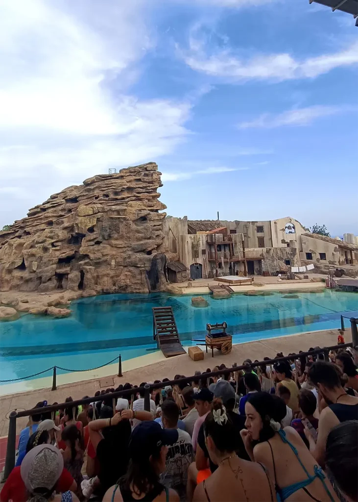 The audience watching a show at Terra Mítica's grand amphitheatre, with the water stage and port town scenery in the background.
