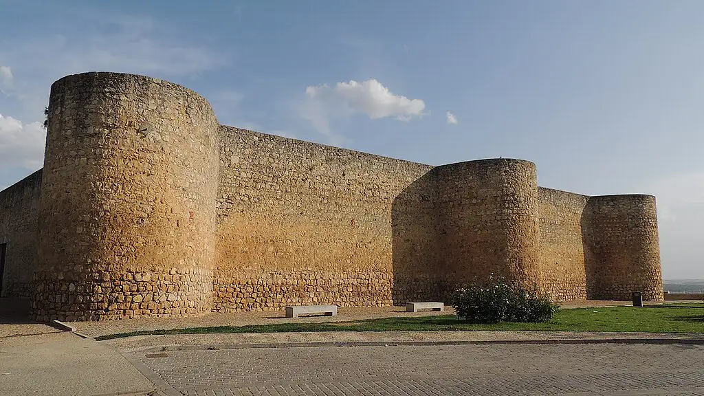 Fachada de pedra do Alcázar de Toro, um antigo castelo real com torres cilíndricas sob um céu azul claro com nuvens, destacando sua arquitetura defensiva.