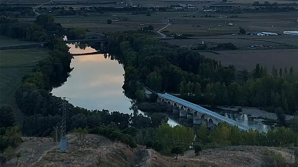 Vista aérea da Ponte Medieval de Toro com seus múltiplos arcos de pedra sobre o rio Douro, rodeada por árvores e campos, ao entardecer.