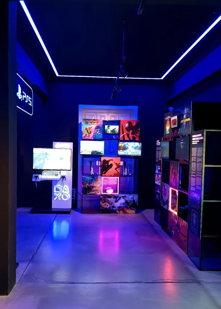 Sala do OXO Madrid com monitores e jogos do PlayStation 5 em exposição, decorada com luzes de neon azuis e roxas.