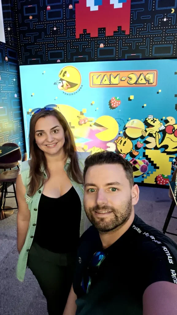 Casal sorrindo em frente ao painel do Pac-Man no OXO Madrid, com cenário comemorativo do 45º aniversário.