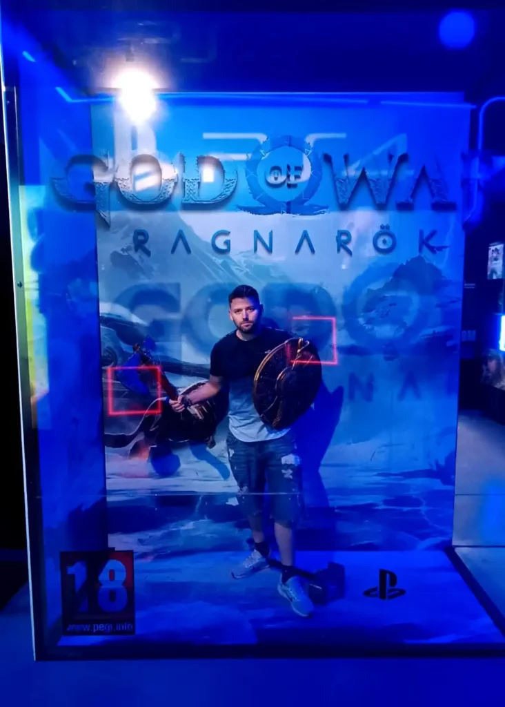 Visitante dentro de cenário interativo de God of War Ragnarök com armas e escudo.