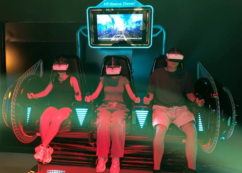 Três pessoas sentadas em simulador de realidade virtual no OXO Madrid, usando óculos VR e controles.