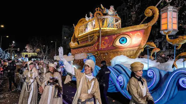 Desfile da Cabalgata de Reyes com figurantes vestidos com trajes inspirados no Oriente, caminhando ao lado de uma carroça iluminada em forma de barco, durante a celebração de 5 de janeiro na Espanha.