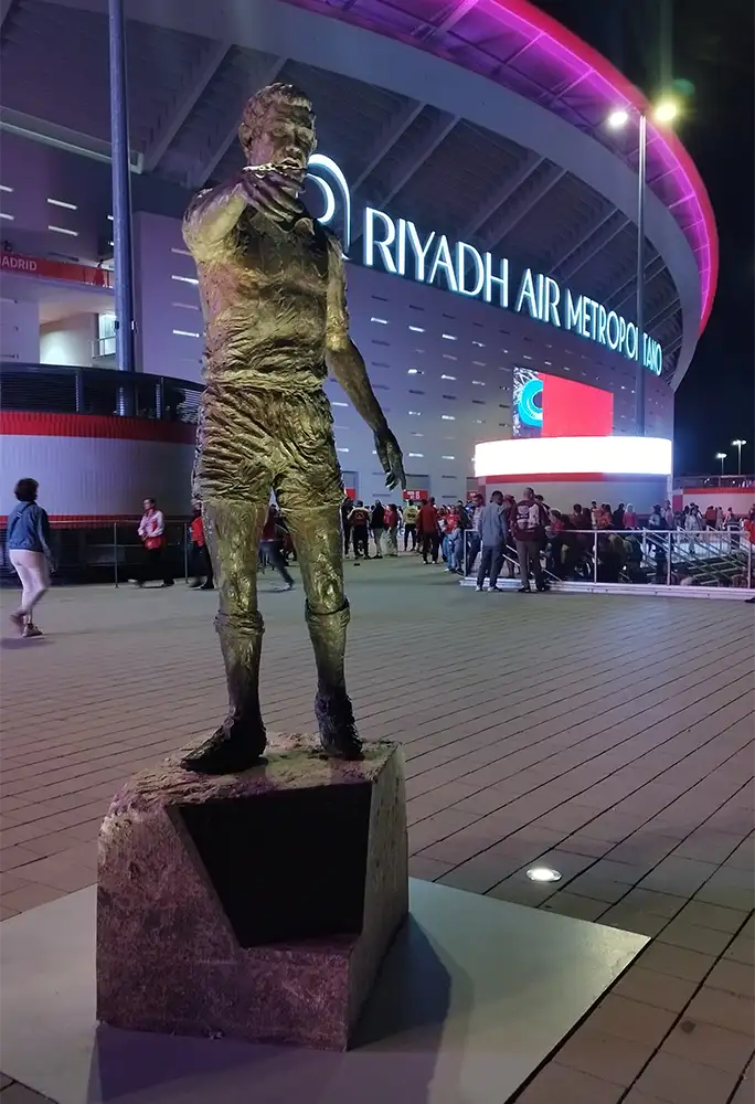 Estátua de jogador de futebol diante do Estádio Metropolitano de Madrid iluminado em tons de rosa.