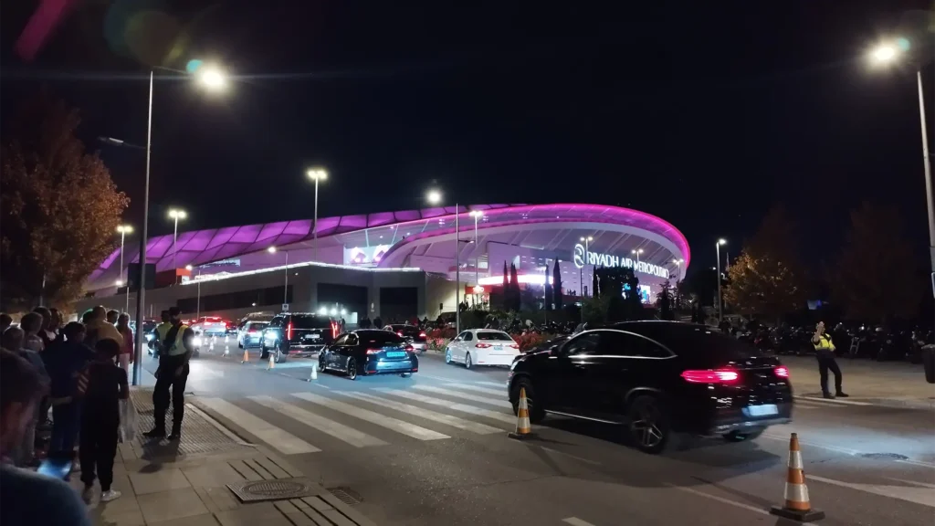 Entrada iluminada do Estádio Cívitas Metropolitano em Madri à noite, com carros e pessoas chegando para um jogo.