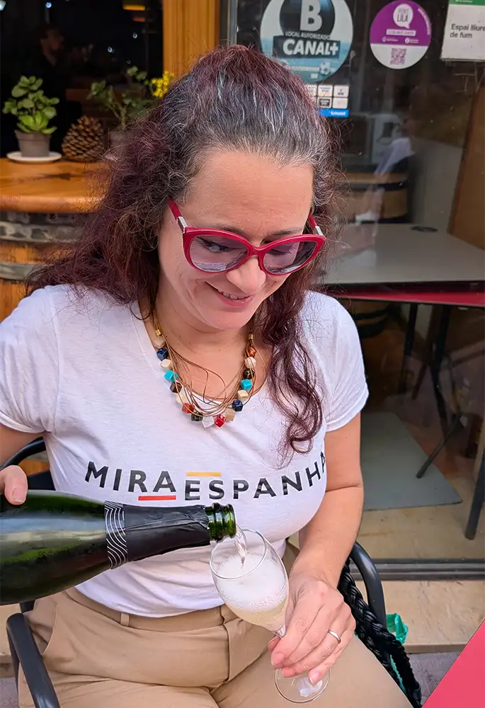 Mulher sentada em um terraço servindo cava em uma taça, usando camiseta do Mira Espanha, durante visita ao Penedès.