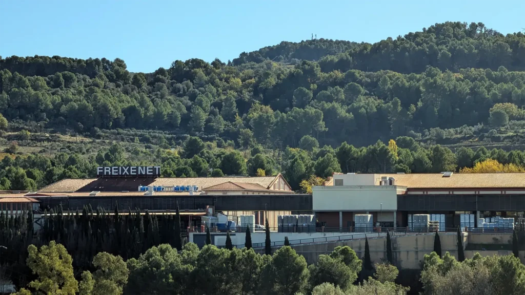 Vista das instalações da Freixenet no Penedès, rodeadas por colinas cobertas de vegetação e vinhas, com edifícios industriais ao pé da serra.