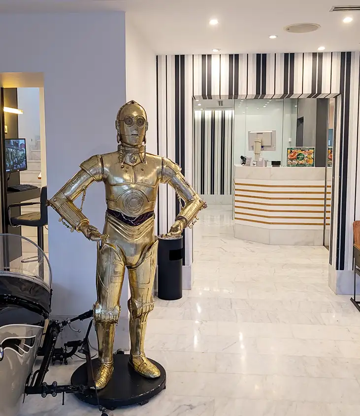 Área interna do Hotel Sitges com recepção ao fundo e estátua do C-3PO em destaque no espaço comum.