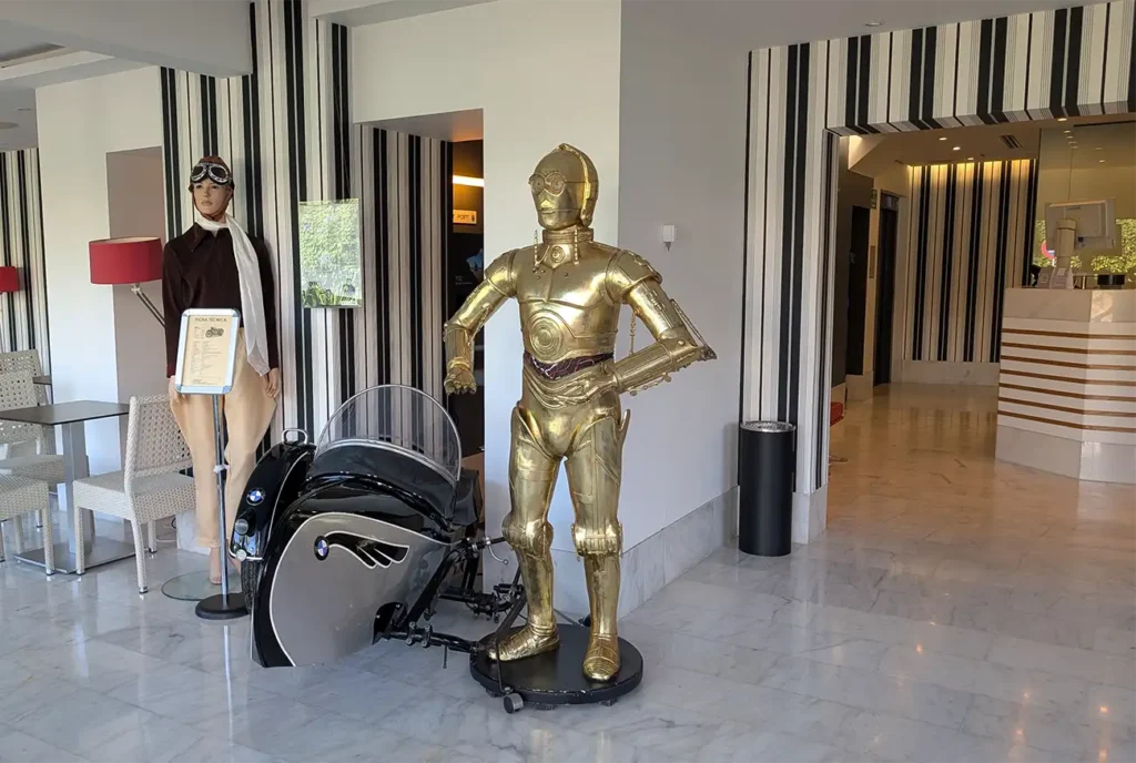 Estátua do personagem C-3PO ao lado de uma motocicleta no lobby do Hotel Sitges, com decoração moderna ao fundo.