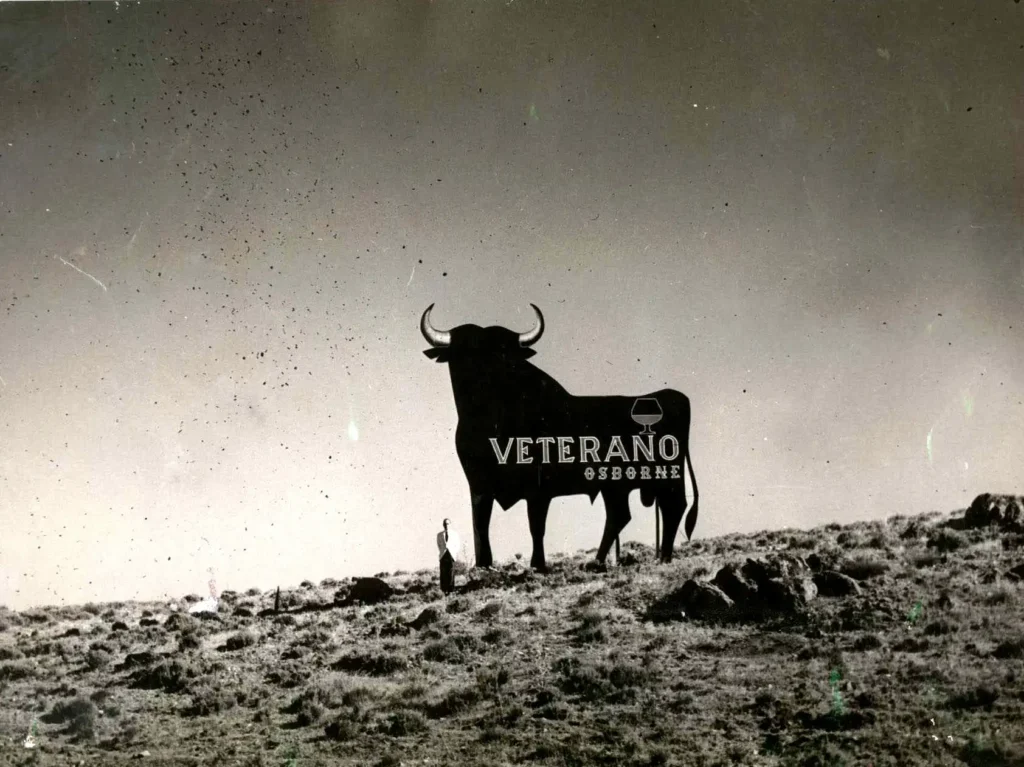Fotografia histórica do primeiro Touro de Osborne com a palavra Veterano e o nome Osborne visíveis na estrutura, instalado num terreno rural, com uma pessoa ao lado para escala.