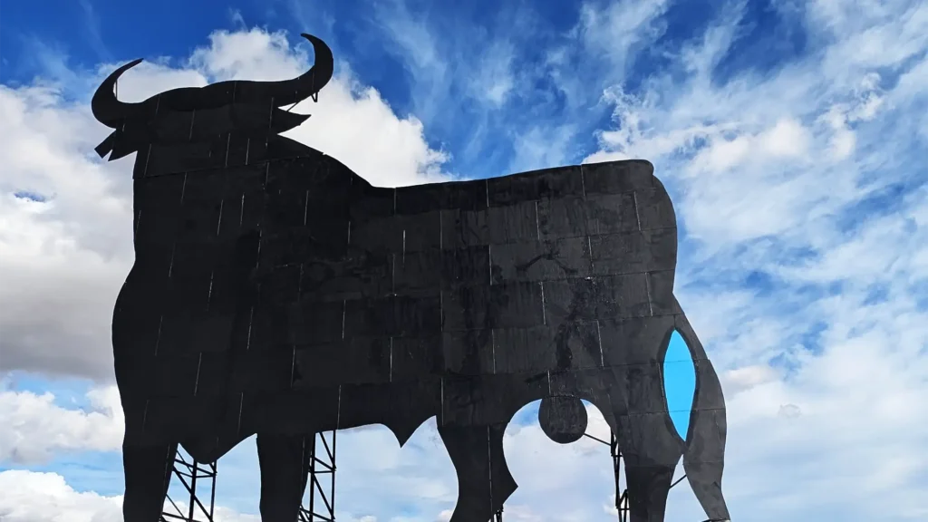 Silhueta do Touro de Osborne nos Monegros, estrutura metálica preta recortada contra o céu azul com nuvens, símbolo cultural da Espanha.