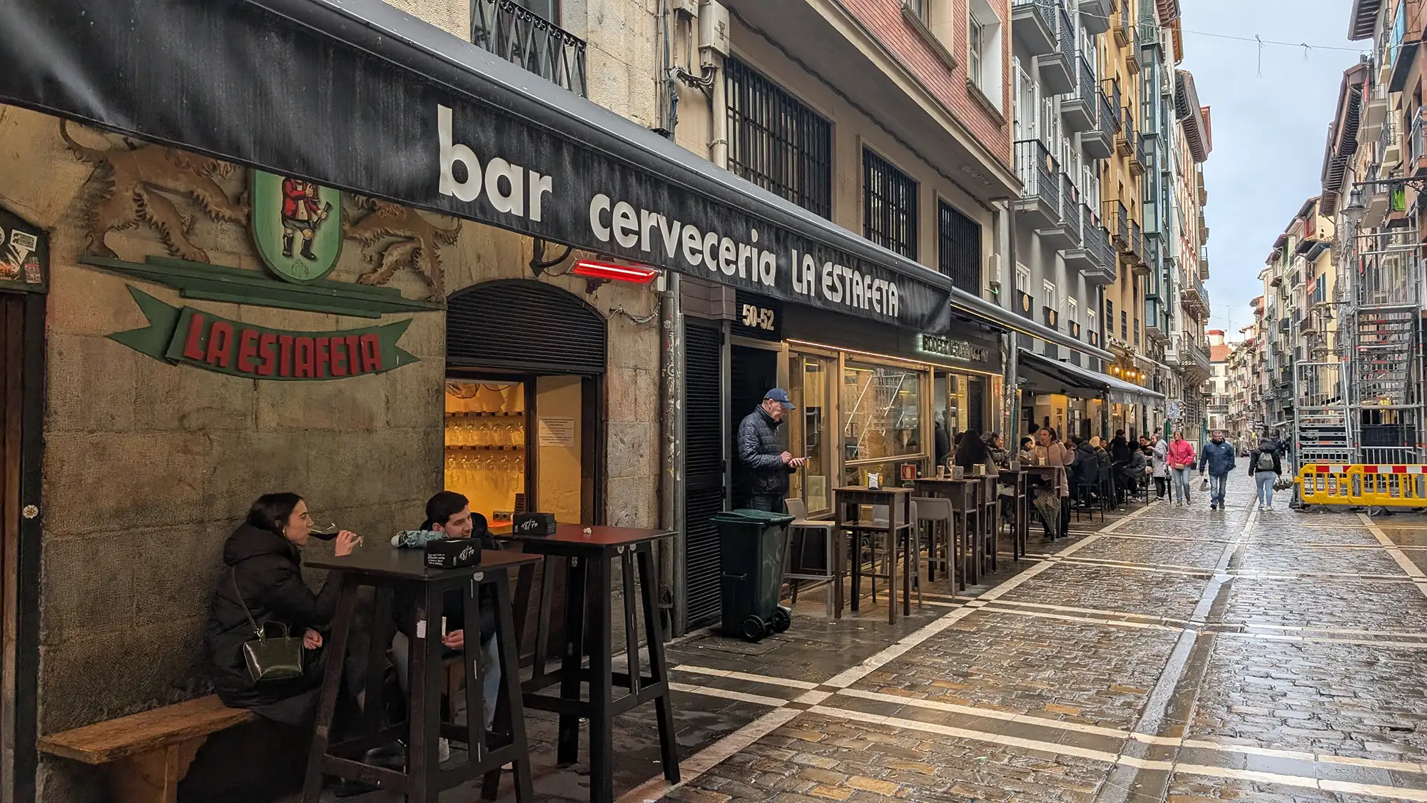 Bar La Estafeta com mesas na calçada da Calle Estafeta, em Pamplona, com pessoas sentadas e prédios históricos ao fundo.