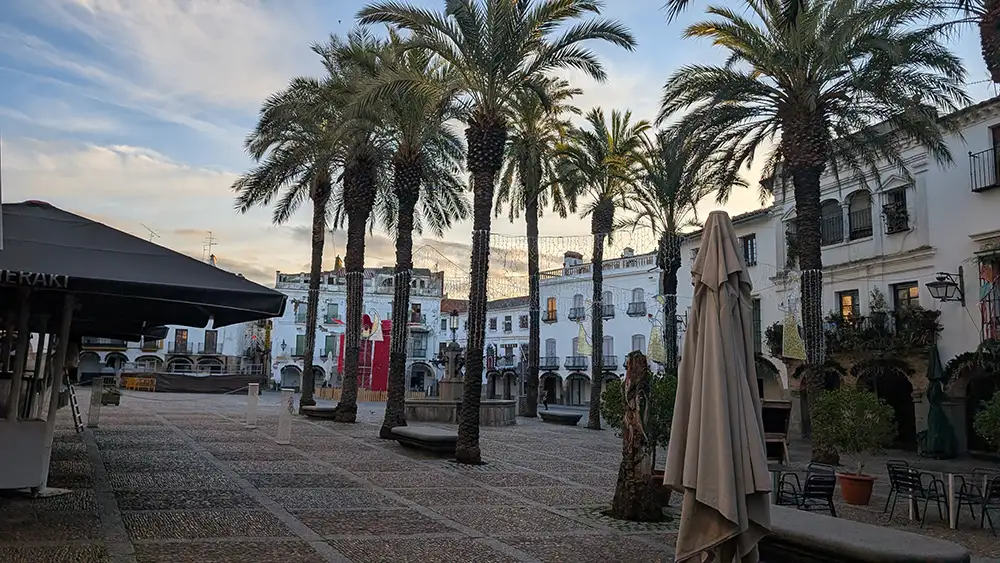 Plaza Grande de Zafra com palmeiras alinhadas, edifícios históricos ao redor e mesas de bares espalhadas pela praça ao entardecer.
