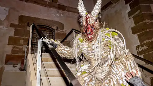 Figura mascarada do carnaval de Águilas com traje tradicional, orelhas longas e máscara expressiva, descendo escadas de pedra à noite.