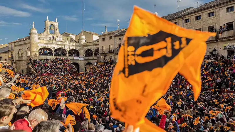 Multidão reunida na Plaza Mayor de Ciudad Rodrigo durante o Carnaval del Toro, com bandeiras laranja erguidas e arquibancadas cheias.