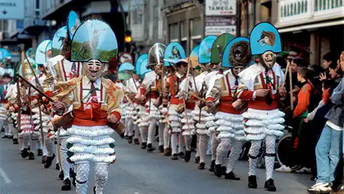 Desfile do Entroido na Galiza com participantes vestidos de branco, máscaras tradicionais e chapéus coloridos, caminhando pelas ruas da cidade.