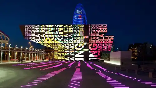 Instalação luminosa do festival Llum Barcelona projetada em edifício moderno, com luzes coloridas iluminando o espaço urbano à noite.