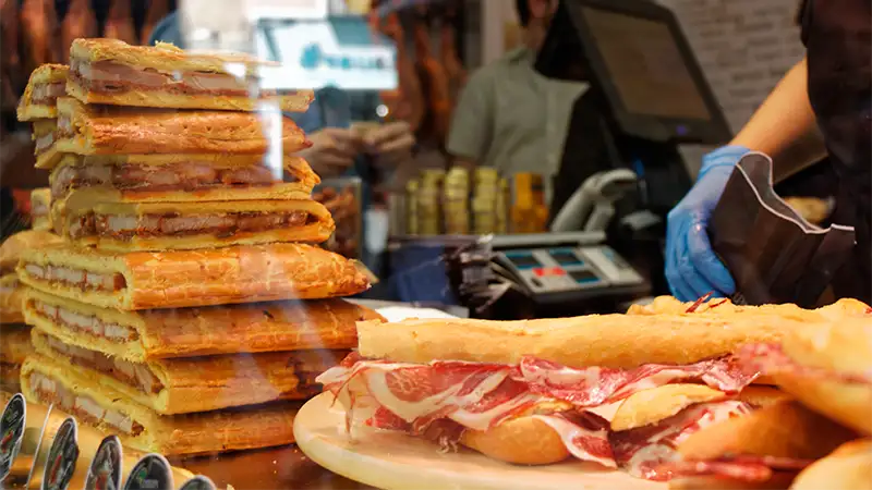 Ham and cheese sandwiches stacked at a market stall in Madrid, with a fresh baguette being prepared behind the counter.