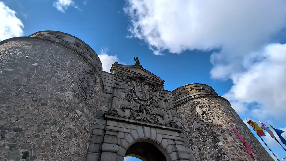 Puerta de Bisagra em Toledo vista de baixo, com torres de pedra e céu azul ao fundo.
