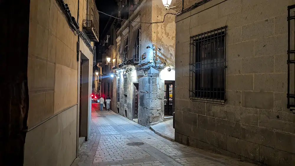 Rua estreita no centro histórico de Toledo à noite com iluminação amarela e fachadas de pedra.