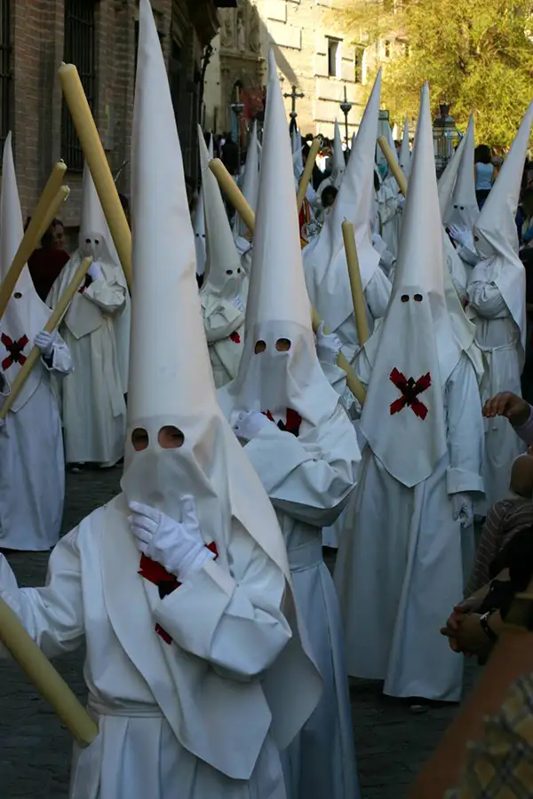 Grupo de nazarenos com túnicas brancas e capuzes pontiagudos carregando velas grandes durante uma procissão de Semana Santa.