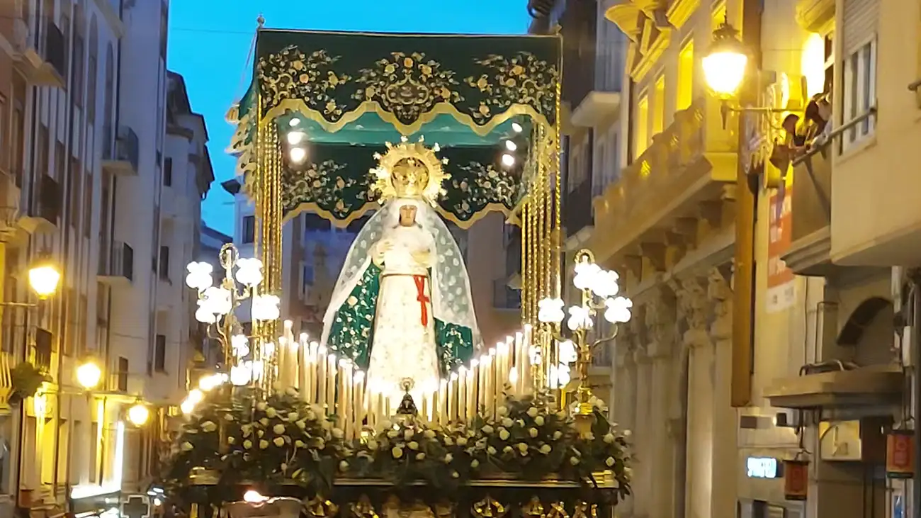 Andor da Virgem Maria em uma procissão de Semana Santa, coberto por um pálio verde e dourado, iluminado por dezenas de velas brancas enquanto passa por uma rua da cidade ao anoitecer.