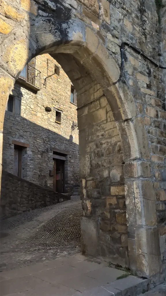 Arco de pedra medieval na entrada do centro histórico de Aínsa com ruela de paralelepípedos e casas de pedra ao fundo.