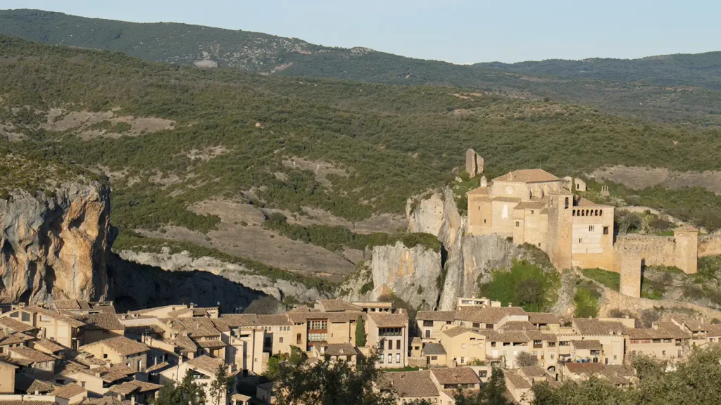 Vista panorâmica de Alquézar, village medieval da província de Huesca, com a colegiata e as muralhas do castelo no alto da rocha e as casas de pedra dourada espalhadas pelo vale, rodeadas por montanhas verdes sob um céu azul claro.