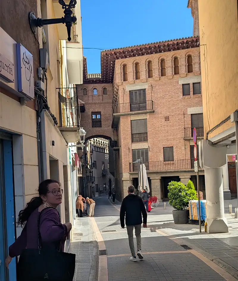 Rua do centro histórico de Graus com edifício de tijolos e arcos mouriscos ao fundo e dois visitantes caminhando, Huesca, Aragão.