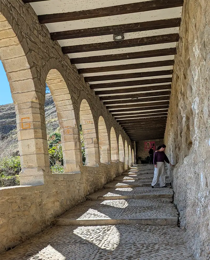 Corredor de arcos de pedra com vigas de madeira no teto do Santuario de la Virgen de la Peña em Graus, com uma pessoa caminhando ao fundo, Huesca.