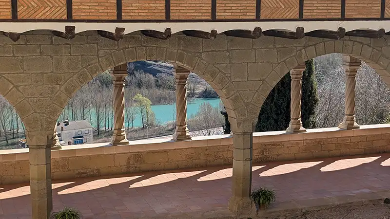 Galeria de arcos de pedra do Santuario de la Virgen de la Peña em Graus com vista para um lago de água turquesa ao fundo, Huesca.
