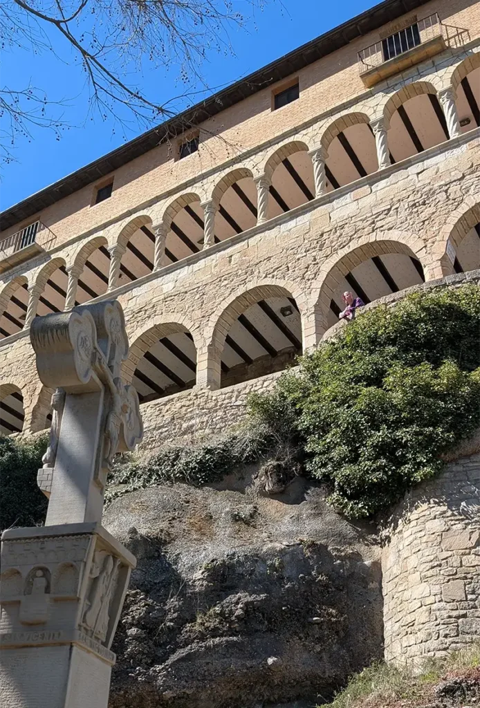 Fachada do Santuario de la Virgen de la Peña em Graus com galerias de arcos em pedra e uma pessoa sentada na rocha abaixo, Huesca