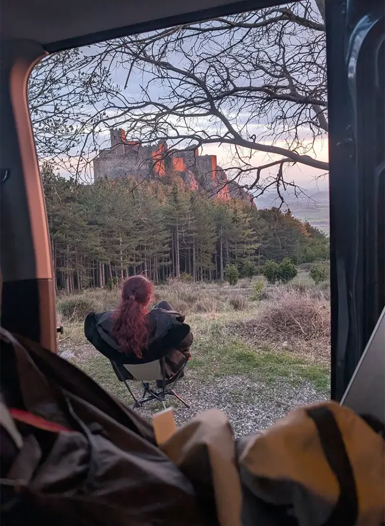 Mulher sentada numa cadeira de camping na porta da van observando o Castillo de Loarre ao entardecer, com o castelo iluminado entre os pinheiros, Huesca, Aragão.