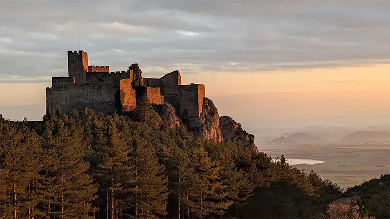Vista panorâmica do Castillo de Loarre no alto da rocha ao pôr do sol, com pinheiros em primeiro plano e o vale de Huesca ao fundo, Aragão.