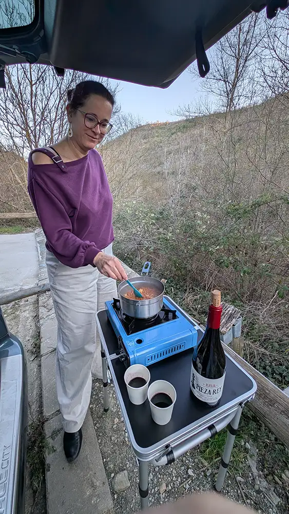 Mulher cozinhando num fogão portátil ao lado da van no estacionamento de Montañana, com uma garrafa de vinho Rioja na mesa, Huesca