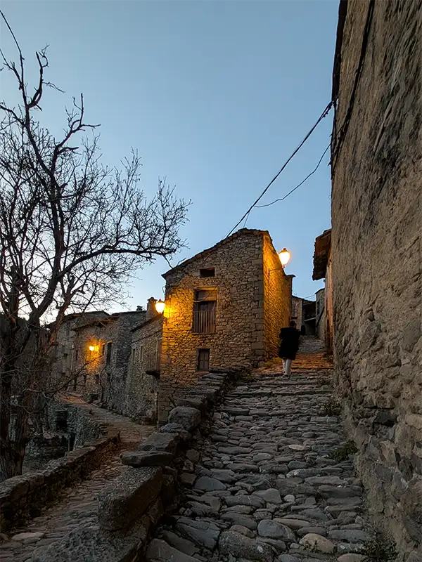 Ruela de pedra de Montañana iluminada por lanternas ao entardecer com casas medievais semiabandonadas e uma pessoa subindo os degraus, Huesca.