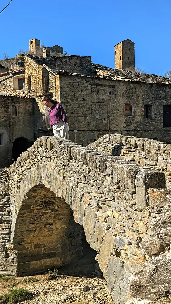 Mulher em pé na ponte medieval de pedra de Montañana com as torres e casas de pedra do povoado semiabandonado ao fundo, Huesca, Aragão.