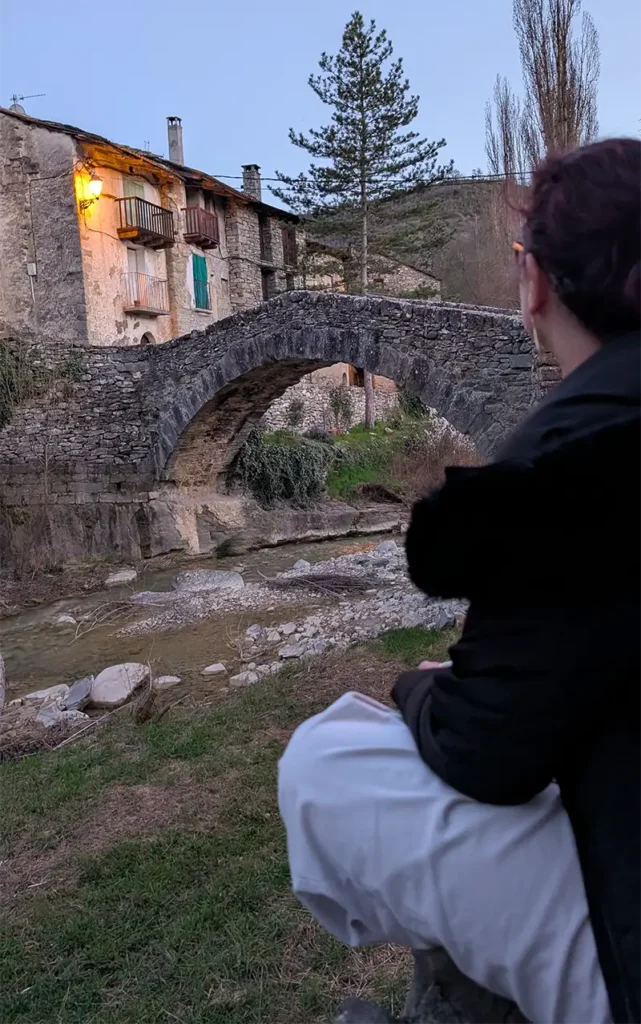 Pessoa sentada observando a ponte medieval de pedra de Montañana ao entardecer com o rio e casas iluminadas ao fundo, Huesca, Aragão.