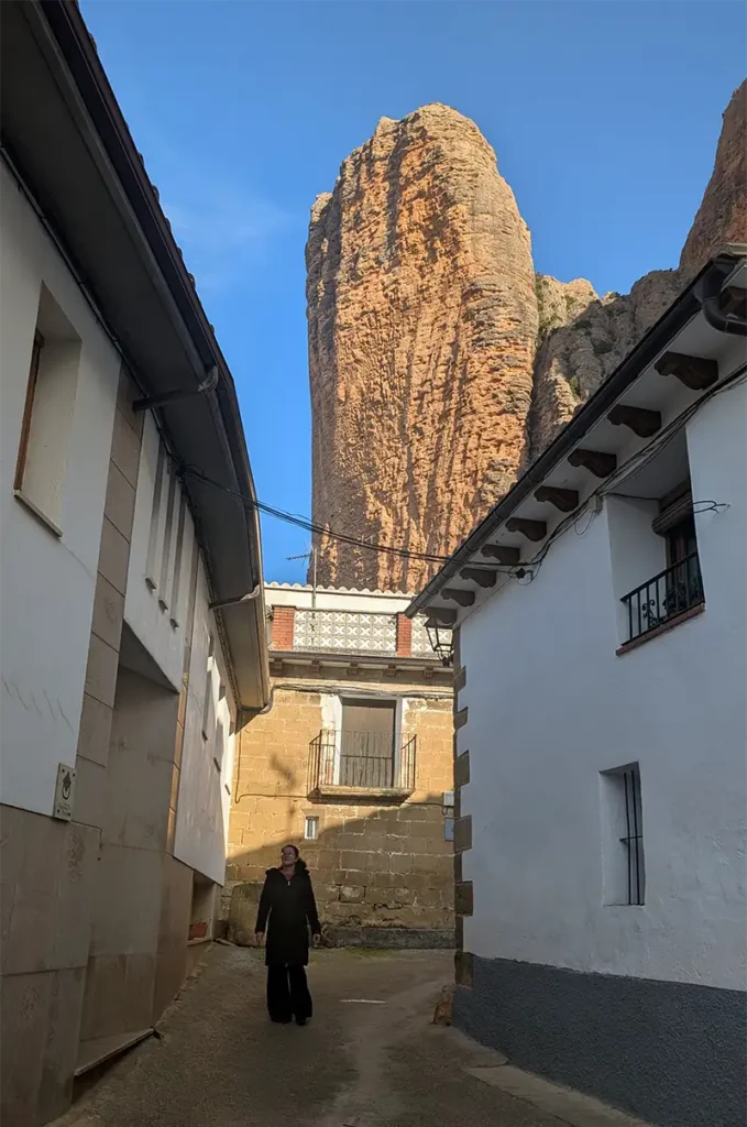 Mulher caminhando por uma ruela estreita de Riglos com um dos mallos de rocha vermelha emergindo entre as casas ao fundo, Huesca, Aragão.